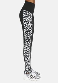 Czarne legginsy z białym wzorem lamparta na bocznym panelu, wysokim pasem i gładką fakturą; w połączeniu z szaro-białymi butami sportowymi.
