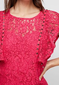 Rosa spetsblus med blommönster, korta ärmar med ruffled detaljer och nitar längs sidorna. Invecklad textur synlig.