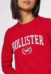 Sudadera roja con texto y logotipo blanco de "HOLLISTER". Cuenta con cuello redondo, puños acanalados y una textura suave y gruesa.