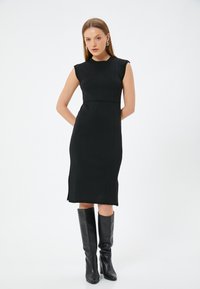 Schwarzes geripptes Kleid mit einer taillierten Silhouette, kurzen Ärmeln und seitlichen Schlitzen. Kombiniert mit knielangen schwarzen Stiefeln für einen eleganten Look.