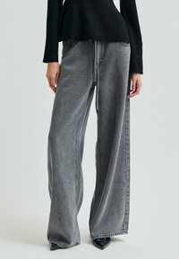 Person som bär svarta spetsiga skor, vida grå denimbyxor med söm fram, och en svart långärmad topp framför en enfärgad bakgrund.