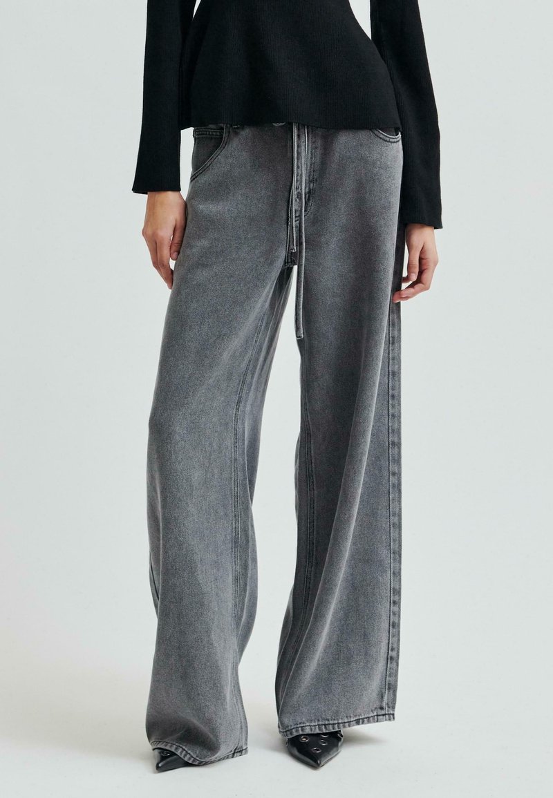 Henkilö, joka on pukeutunut mustiin kärkitossuihin, leveisiin harmaisiin farkkuihin, joissa on etusaumadetaili, ja mustaan pitkähihaiseen paitaan yksinkertaista taustaa vasten.