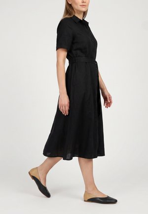 Frau in schwarzem Midi-Kleid mit kurzen Ärmeln, Knopfleiste vorne und Gürtel, trägt zweifarbige schwarze und beige flache Schuhe, geht vor einfarbigem Hintergrund.