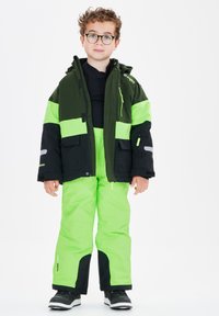Giacca impermeabile verde e nera con accenti riflettenti e pantaloni abbinati di un brillante verde. Capelli ricci, occhiali e maglietta nera. Visibili stivali da neve.