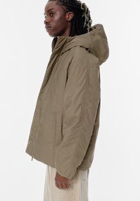 Kurtka z kapturem w stylu beanie w kolorze khaki, z zamkiem z przodu, bocznymi kieszeniami i teksturowanym materiałem. Charakteryzuje się luźnym krojem i bezszwowym wykończeniem ramion.