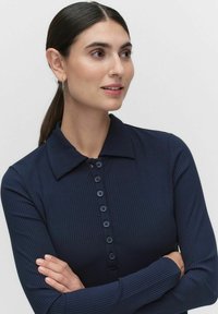 Laivan sininen ribattua pitkähihaista paitoa kauluksella, jossa on napitettava etuosa ja teksturoitu kangas. Suunnitelma sisältää hienovaraisen neulostyylin.