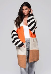 Przeciętny kardigan w kolorach pomarańczowym, kremowym i brązowym z czarno-białymi paskami. Dzianinowa faktura, otwarty przód i szerokie rękawy.