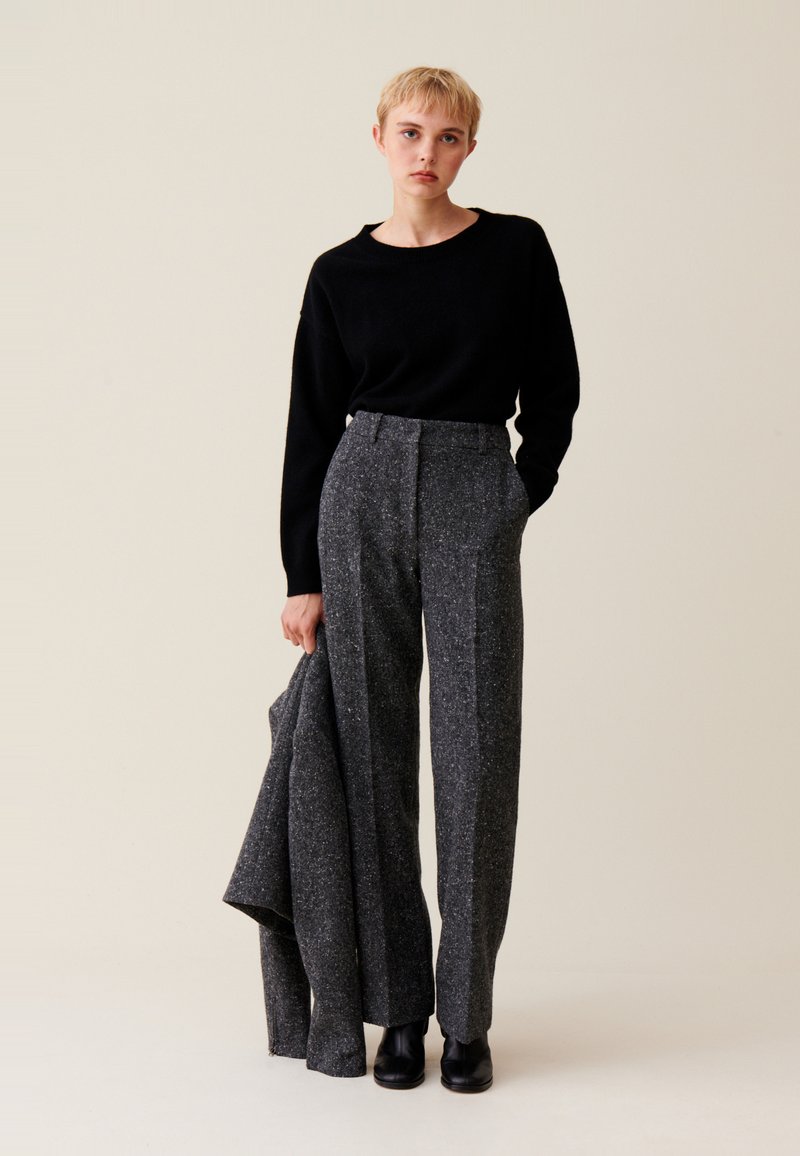Czarny sweter zestawiony z wysokogatunkowymi, szerokimi spodniami z szarej tkaniny tweed, z subtelną fakturą i ukrytymi kieszeniami. Modelka trzyma pasującą kurtkę.