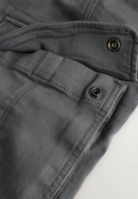 Sive bombažne hlače z mehko teksturo, vidnimi šivi in črnim kovinskim okovjem na žepih. Prikaz v bližnjem posnetku, ki poudarja podrobnosti.