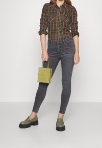 Camicia a quadri verde e rossa, jeans skinny grigi, mocassini verde oliva e una piccola borsa verde testurizzata. Sfondo a superficie piana.