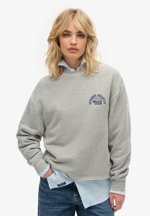 Grijze sweatshirt met een ronde hals, oversized pasvorm en blauwe opdruk 'Superdry Athletics Beach Club'. Gecombineerd met een lichtblauw gestreefd shirt eronder.