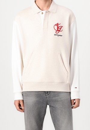 Beige pullover med hvide lange ærmer, der har en forlomme, broderet rød og sort logo samt ribstrikkede manchetter.