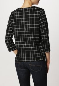 Žena oblečená v černobílém kostkovaném tričku s dlouhým rukávem a zadním zipem, v kombinaci s tmavě modrými džínami, zobrazena zezadu.