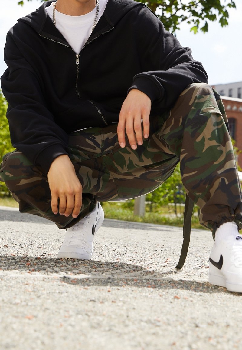 Sudadera negra con cremallera, pantalones cargo de camuflaje y zapatillas blancas con detalles negros. Pose en cuclillas sobre una superficie de grava. Árboles verdes al fondo.