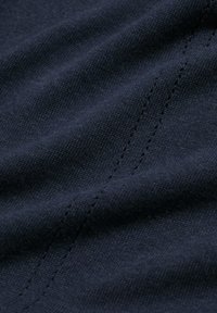 Lähikuva tummansinisestä kankaasta, jossa on hieno rakenne ja kaksi rinnakkaista riviä pieniä, siistejä pistoja, jotka kulkevat vinottain pinnan yli.