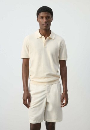 Joven de pie vestido con una camiseta polo crema de manga corta y pantalones cortos crema a juego sobre un fondo liso claro.