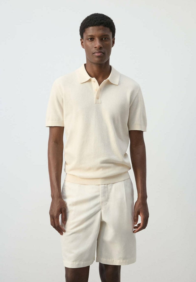 Jeune homme debout portant un polo crème à manches courtes et un short crème assorti devant un fond clair uni.