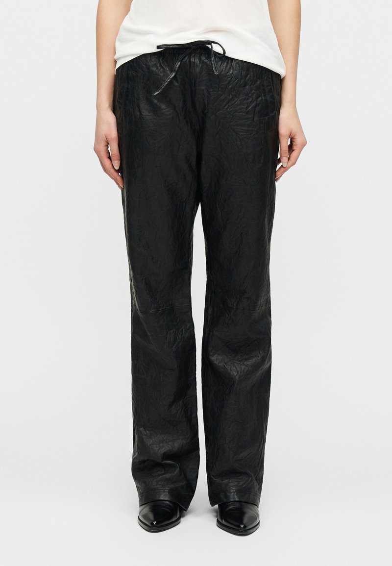 Άτομο με λευκό πουκάμισο που φοράει μαύρο υφασμάτινο παντελόνι με ίσια πόδια και κορδόνι στη μέση, καθώς και μαύρα παπούτσια με μυτερή μύτη, στέκεται σε λευκό φόντο.