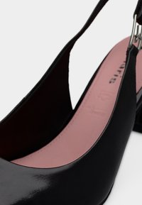 Gros plan sur une chaussure slingback en cuir noir avec une semelle intérieure rose et une boucle argentée sur fond blanc.