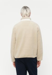 Beige fleece genser med høy krage, teksturert overflate og avslappet passform. Har en subtil søm på den øvre delen av ryggen.
