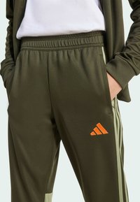 Maslinasto zelene sportske hlače s teksturiranim završetkom, s narandžastim Adidas logotipom i svijetlozelenim bočnim trakama. Elastična gumica u struku i džepovi.