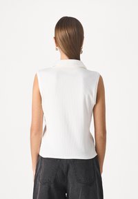 Femme avec des cheveux bruns raides portant un haut sans manches blanc et un jean noir taille haute, vue de dos sur fond blanc.