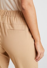 Pantalons beiges extensibles avec une taille élastique et une seule poche arrière. Le tissu est lisse avec une texture douce.