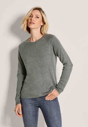 Vrouw met blond haar die een grijze trui met lange mouwen en patroonmouwen draagt, en een blauwe spijkerbroek, staand tegen een effen achtergrond.