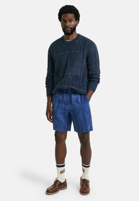 Maglione a maglia navy con motivo testurizzato, abbinato a pantaloni corti blu, calze bianche con striscia rossa e scritte, e scarpe marroni.
