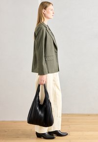 Must mustriga mustekott musta pleedi tekstuuriga, mille hoiab käes inimene oliivrohelises triibulises bleiseris ja kreemivärvi laia jalaga pükstes.