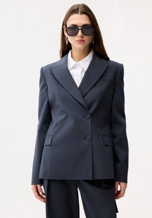 Kobieta ubrana w ciemny, teksturowany, oversize'owy żakiet oraz pasujące spodnie, białą koszulę z kołnierzykiem, oversize'owe czarne okulary przeciwsłoneczne i srebrne kolczyki-koła.