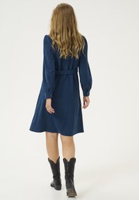 Námornícky modré kordurové šaty s dlhými rukávmi, opaskom v páse a sukňou po kolená. Nosené s čiernymi kovbojskými čižmami.