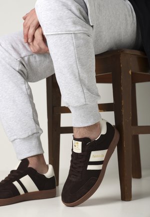 Sneaker in scamosciato marrone con dettagli bianchi e strisce nere. Pantaloni della tuta in cotone grigio con polsini a costine. Sedia di legno sullo sfondo.