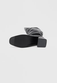 Botte à talon noir avec une tige lisse et brillante, un bout carré et une semelle en caoutchouc texturé. Présente un design froncé pour un effet drapé.