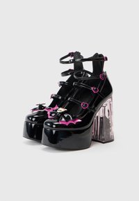 Pantofi platform cu aspect patent negru, cu accente roz sub forma de lilieci și cranii, având mai multe curele și un toc bloc transparent. Textură netedă pe toată suprafața.