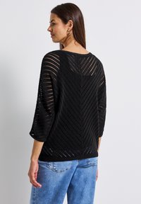 Schwarzer Strickpullover mit Zickzackmuster, Dreiviertelärmeln und lockerer Passform, kombiniert mit hellblauen Jeans.