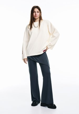 Roomwit oversized sweatshirt met lange mouwen, gecombineerd met donkergrijze asymmetrische broek en zwarte plateauzolen, met een subtiel logodetail.