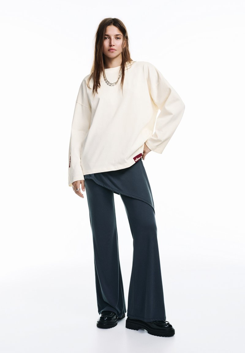 Krém színű, bő szabású pulóver hosszú ujjakkal, sötétszürke aszimmetrikus nadrággal és fekete platform cipővel, finom logó részlettel.