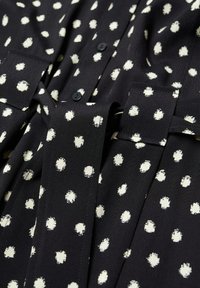 Blouse en tissu noir ornée de pois blancs de forme irrégulière, avec des fermetures à boutons et un détail de taille nouée. Matière douce et texturée.