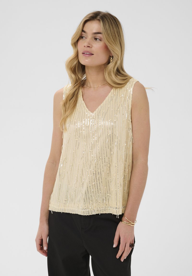 Femme aux longs cheveux blonds portant un haut beige à sequins sans manches et un pantalon noir, regardant légèrement vers la gauche sur un fond uni.