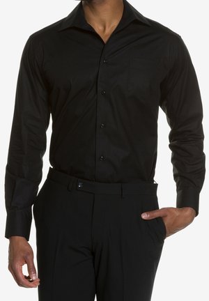 Mężczyzna w dopasowanej czarnej koszuli wizytowej włożonej do czarnych spodni wizytowych, z jedną ręką w kieszeni, na tle białego tła.