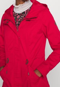 Veste rouge à capuche avec fermetures éclair et à boutons, taille ajustable par cordon de serrage, et deux poches avant. Fabriquée en tissu lisse avec une finition mate.