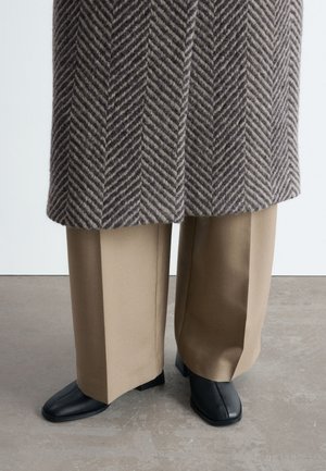 Jambes inférieures en pantalon beige à jambes larges, chaussures noires à bout carré, et un manteau gris et marron à motif chevrons. Debout sur un sol gris.