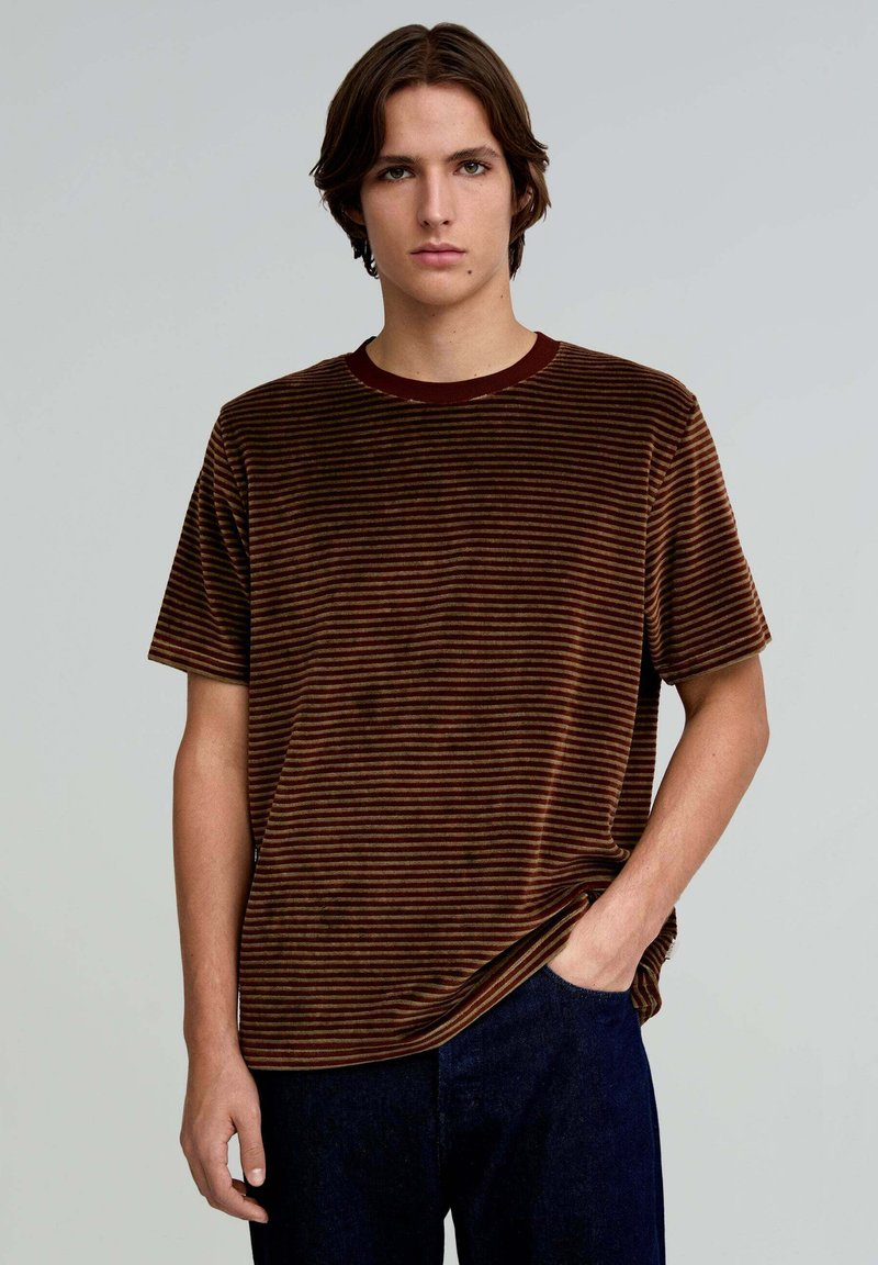 Brunstribet kortærmet T-shirt i blødt tekstureret stof, med en bordeaux rund hals og afslappet pasform. Båret med mørkeblå bukser.