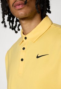 Κίτρινο πόλο μπλουζάκι με μαλακή υφή, διαθέτοντας μαύρο λογότυπο Nike, τρία μαύρα κουμπιά στη λαιμόκοψη και σχέδιο για γιακά.