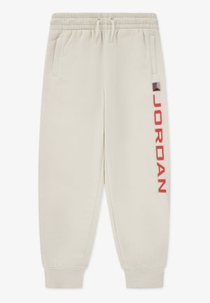 Pantalon de survêtement beige clair avec taille et poignets élastiques, poches latérales, et texte vertical rouge "Jordan" avec logo sur la jambe droite.