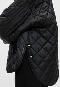 Melna quiltota jaka, izgatavota no gludas auduma, ar spiedpogām un logo tagu, ar plūstošu dizainu, kas uzsver tās siltīgo, pildījuma tekstūru.