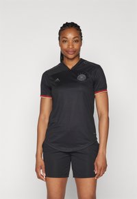 Svart fotbollströja med v-ringning, med röda och gyllene detaljer på ärmarna och en cirkulär logotyp. Bärs med matchande svarta shorts.