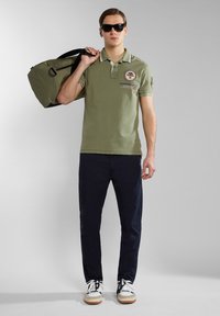 Oliwkowa koszulka polo z kołnierzykiem i haftowanym naszywkiem, w połączeniu z ciemnogranatowymi spodniami i białymi trampkami, niosąca zieloną torbę sportową.