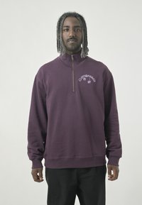 Lila Zip-Up Sweatshirt mit hohem Kragen, mit weißen Druckdetails auf der Brust. Hergestellt aus einem weichen, strukturierten Stoff.